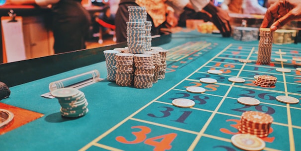 A casino scene with a roulette table features prominently, covered in green felt with colorful numbers and betting squares. Several stacks of poker chips are placed on different numbers, and the hands of several people are visible as they interact with the table. In the background, there are slot machines and more people, creating a vibrant and busy atmosphere.