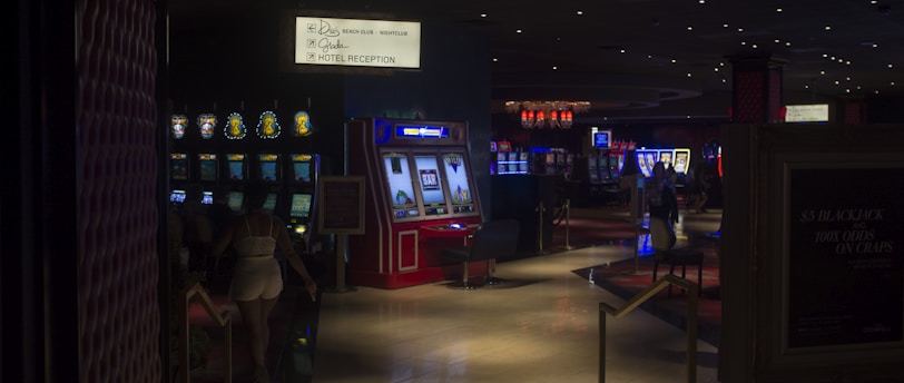 A dimly lit casino with several rows of slot machines, some displaying bright graphics and lights. There are a few people scattered throughout the area, engaging with the machines. The floor is reflective and polished, adding to the ambiance, while the ceiling is dotted with numerous small lights.