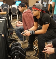 Several people are sitting in a room with gaming consoles and monitors, focusing intently on their screens. The setting appears to be a casual gaming event, with participants holding controllers while playing video games. The room has a casual atmosphere with chairs and tables set up for gaming.