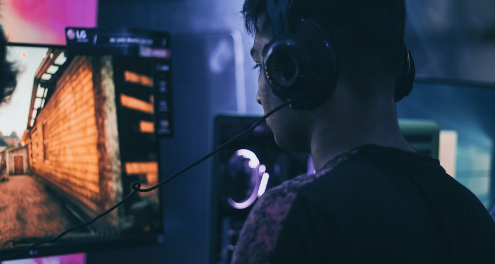 A person wearing headphones is engaged in gaming on a computer, with a large screen displaying a video game scene, showing a wooden building and a path.