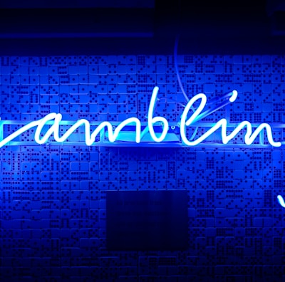 A bright blue neon sign spells out the word 'gambling' in cursive script. The background features a textured wall covered in domino tiles, also illuminated by the blue light, creating a vivid and visually striking scene.