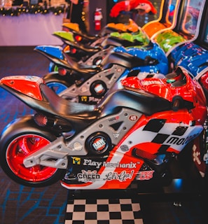 A row of arcade motorcycle racing games is lined up, featuring brightly colored machines with detailed graphics and logos. A person in the background stands near decorated countertops and a wall-mounted screen. The environment has a lively and playful ambiance, with vibrant colors and patterns.