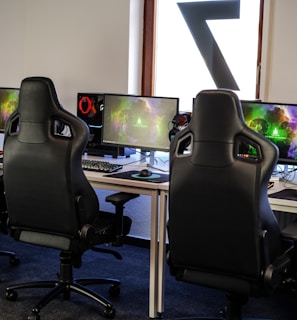 A row of gaming stations equipped with large monitors, gaming chairs, keyboards, and headphones. The setup is organized and ready for gaming, featuring a colorful screen display. The room has a modern and tech-savvy ambiance.