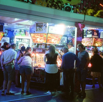 A group of people standing in front of an illuminated amusement arcade, engaging with various gaming machines. The environment is bustling with activity, and colorful lights create a vibrant atmosphere. The ceiling is decorated with large, playful images of dice and other game-related graphics, enhancing the lively mood.