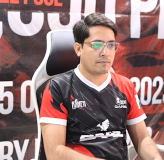 A man with glasses is sitting in a gaming chair, focused on a monitor in front of him. He is wearing a black and red esports jersey with various logos. The background features a large banner with text and numbers in bold fonts.
