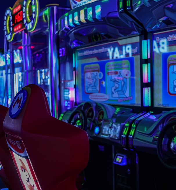 A vibrant arcade environment with several illuminated gaming machines. The image features arcade racing games with visible steering wheels, colorful control panels, and screens displaying game instructions. Bright LED lights in various colors illuminate the machines and surrounding area.